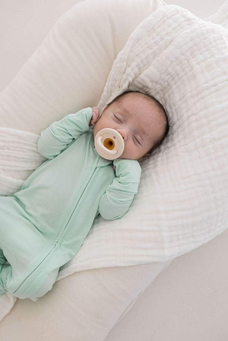 A baby in a mint Bamboo Zip Pajamas by forever french baby rests on a soft white cushion, holding a round beige pacifier. Their hands are near their face as they appear peaceful and content.