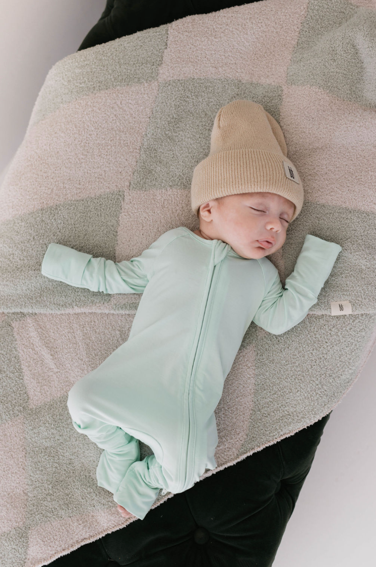 A baby sleeps peacefully on a black surface with a soft, checkered blanket. Wearing breathable Bamboo Zip Pajamas in mint by forever french baby and a beige knit hat, the baby has arms outstretched in blissful slumber.