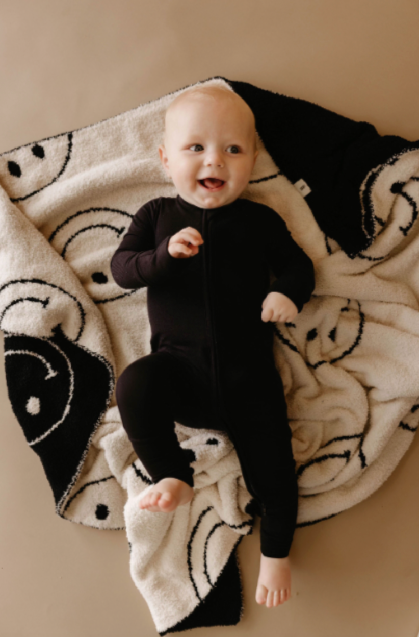 A baby dressed in forever french baby's Bamboo Zip Pajamas in Midnight Black lies on a beige and black blanket adorned with smiley faces. The baby has a cheerful expression and looks off to the side with a smile. The background is a light brown color.