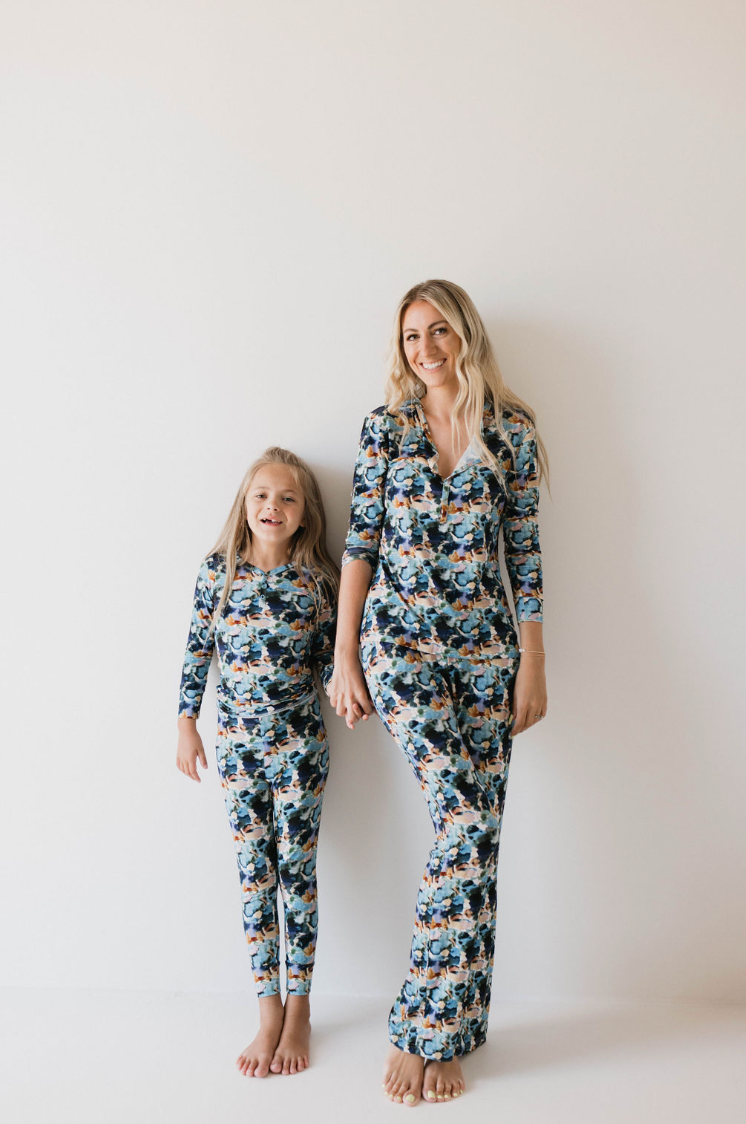 A woman and a young girl stand side by side against a plain white background, smiling and holding hands while wearing matching Bamboo Two Piece Pajamas in the Charli Print from forever french baby. The woman has long blonde hair, and the girl has her long blonde hair tied in a ponytail.