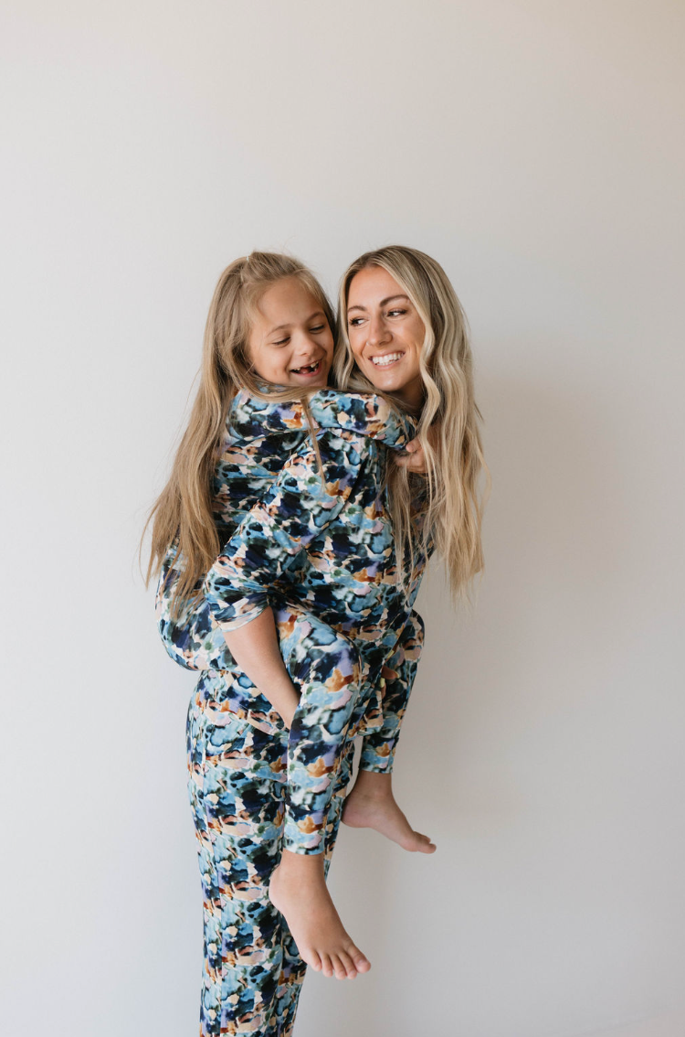 A woman with long blonde hair smiles as she gives a piggyback ride to a young girl with light brown hair. Both are dressed in matching colorful floral outfits from the "Bamboo Two Piece Pajamas | Charli Print" collection by forever french baby, posed against a plain white background to promote autism awareness and the importance of early intervention.