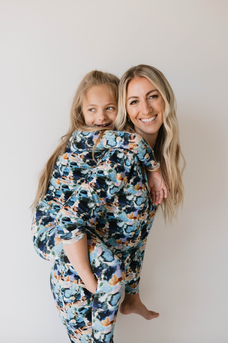 A woman with long blonde hair smiles at the camera while giving a piggyback ride to a young girl with light brown hair. Both are dressed in matching forever french baby's Bamboo Two Piece Pajamas in Charli Print, featuring colorful, abstract patterns that promote autism awareness. They stand against a plain white background.