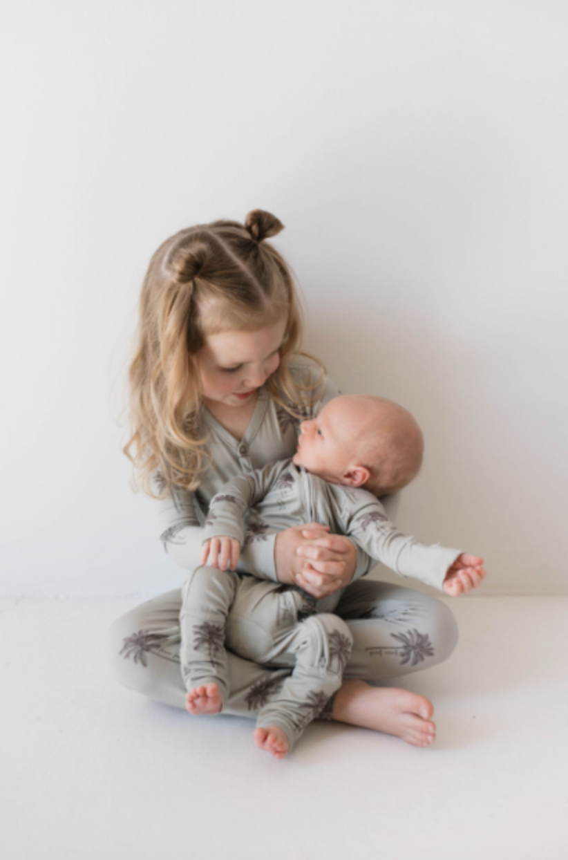 A blonde girl in a gray "Summer Dreamin'" Bamboo Zip Pajamas from forever french baby, featuring dark palm tree prints, holds a baby in matching pajamas. Both children seem content against a plain white background, showcasing the comfort of these pajamas.