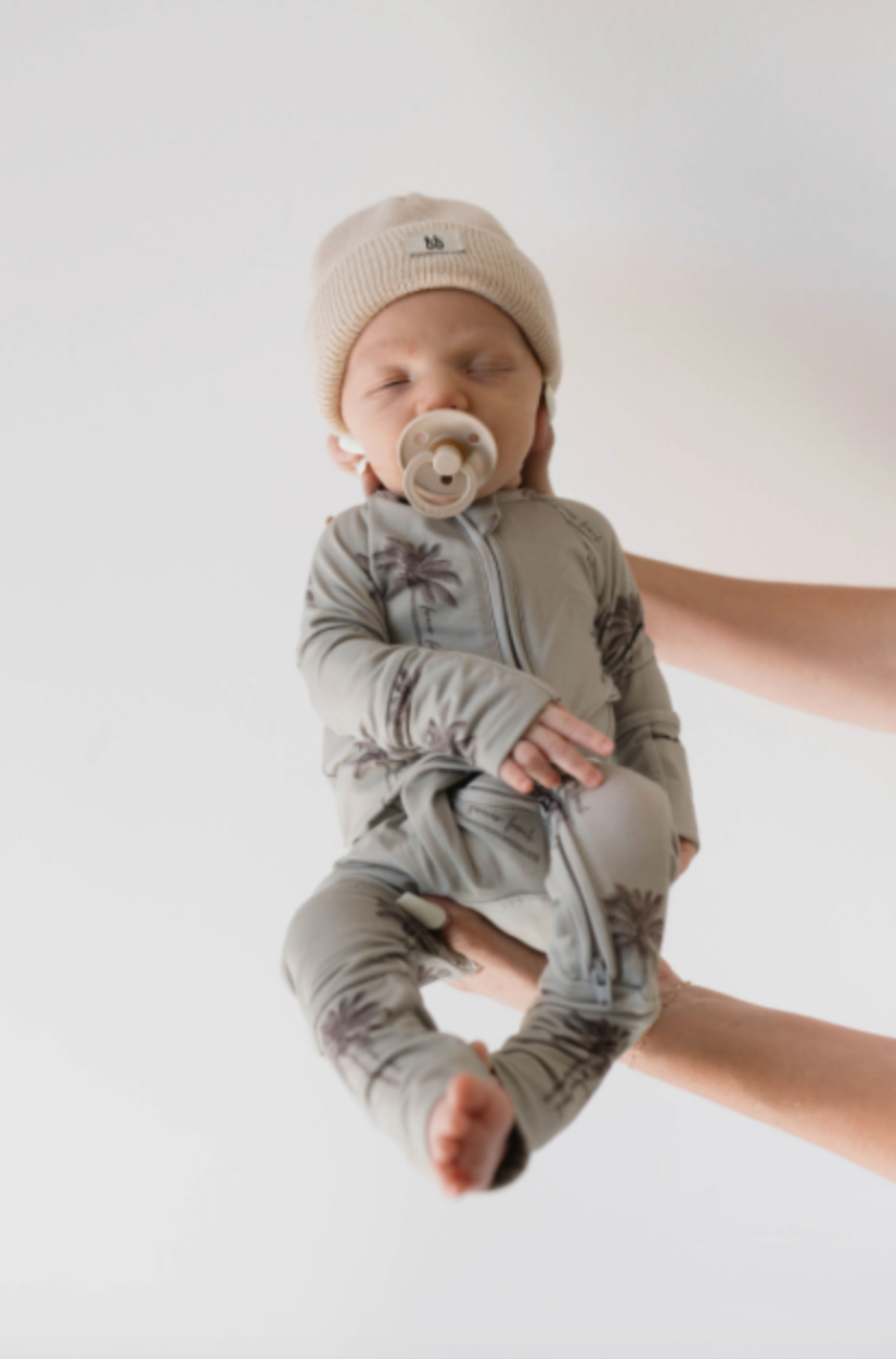 A baby dressed in Forever French Baby's Bamboo Zip Pajamas, Summer Dreamin', is gently held against a plain white backdrop. With a beige knit hat and pacifier, the little one seems to dream peacefully of summer beaches.