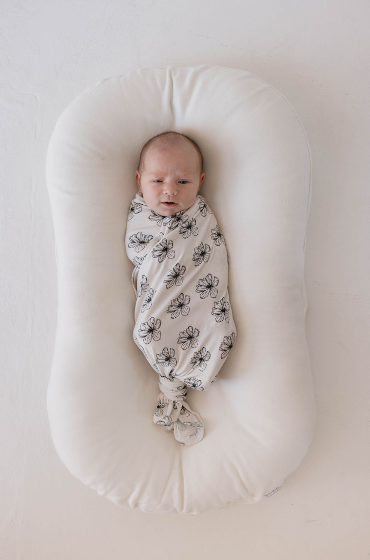 A newborn baby is swaddled in the hypo-allergenic Bamboo Swaddle | Desert Bloom by forever french baby, lying on a white, cushioned surface. The baby has eyes open and is positioned in the center of the image, facing upward. The background is a light, neutral color.