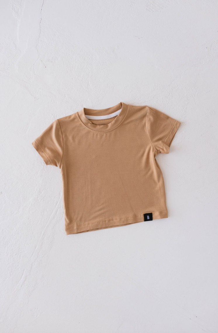 A Basic Bamboo Tee in Desert Sunset from forever french baby is laid flat against a light background. The comfortable, plain, short-sleeved T-shirt is made from soft bamboo fabric and features a small black tag at the bottom hem on the left side.