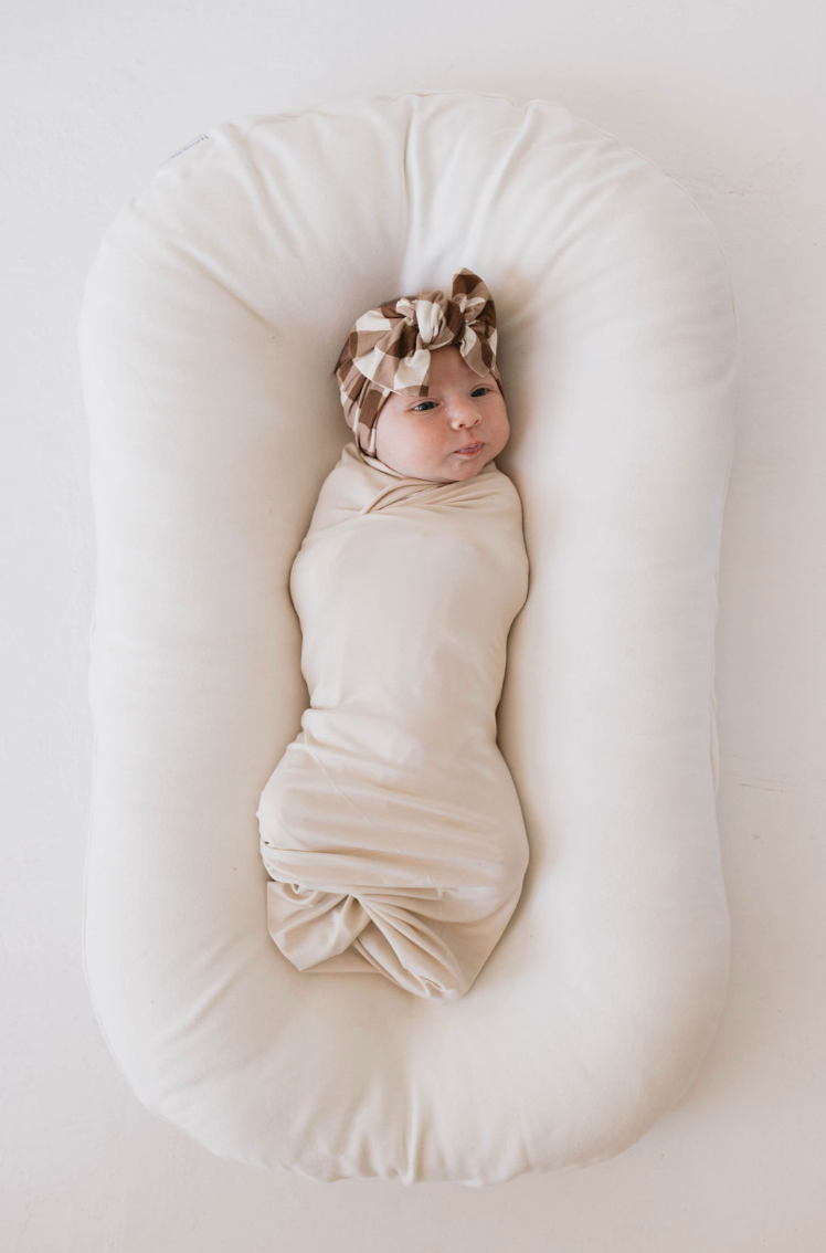 A baby is swaddled in a Bamboo Swaddle | Bone by forever french baby and lying on a soft, oval-shaped white cushion. The baby is wearing a brown and cream checkered headband with a bow, looking up with a content expression. The background is a simple white surface.