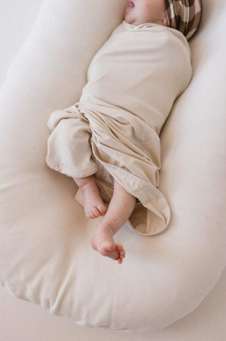 A baby, wrapped in the forever french baby's Bamboo Swaddle in Bone and wearing a matching striped headband, is lying on a soft, white cushion. The baby's legs and feet are partially visible, with one leg extended while the other remains bent. The baby appears to be sleeping peacefully.