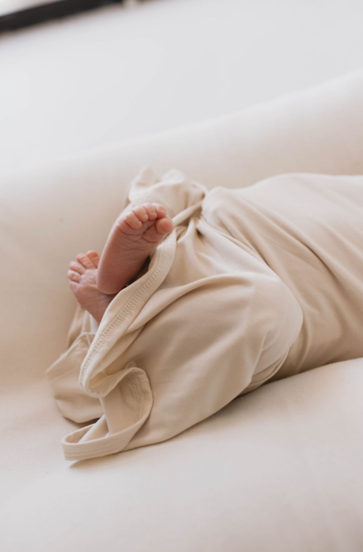 A baby wrapped in a soft, Bone-colored Bamboo Swaddle from forever french baby lies on a light-colored surface. Only the baby's tiny feet peek out from the blanket. The scene is gentle and serene, emphasizing the comfort and tenderness of the moment.