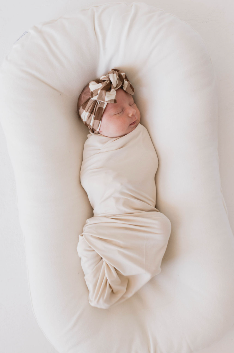 A newborn baby is swaddled in a Bone-colored Bamboo Swaddle from Forever French Baby, lying on a soft, white pillow. The baby is wearing a brown and white patterned headband and is sleeping peacefully. The background is white, creating a serene and minimalist setting.