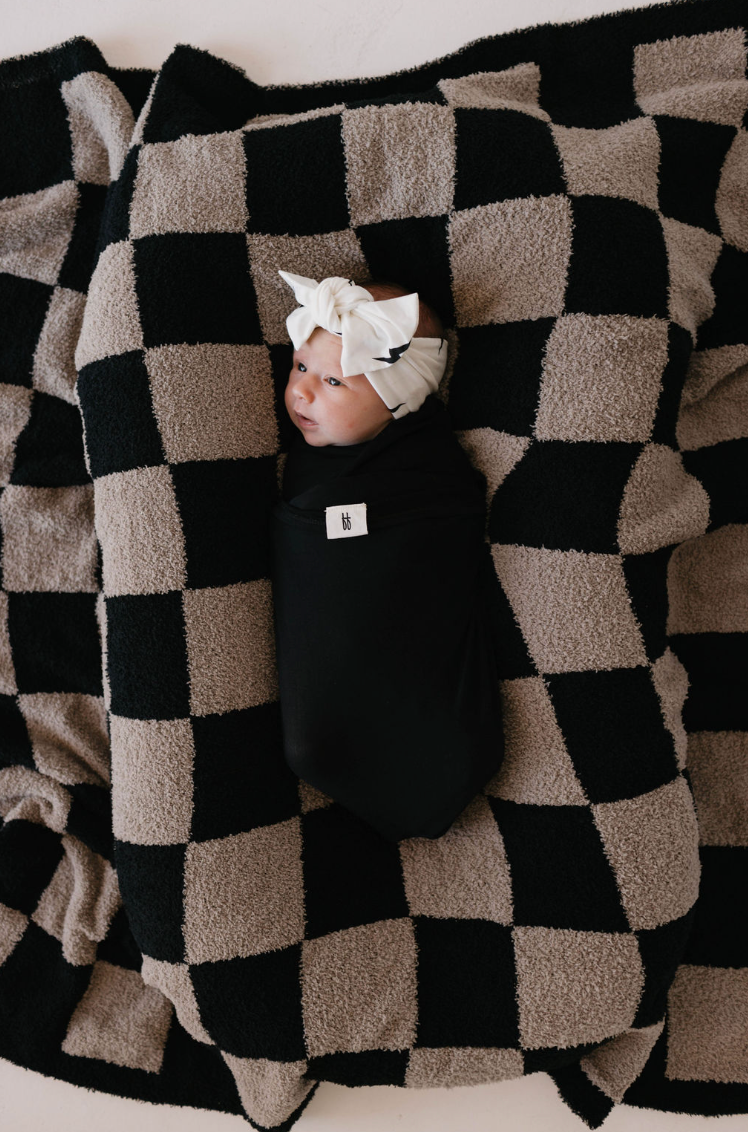 A baby wrapped in a black blanket with a white bow headband is lying on the plush "Checkmate Checkerboard" blanket from forever french baby. This cozy and content little one looks to the side, snuggled in the softness of an Oeko-tex certified fabric for ultimate safety and comfort.