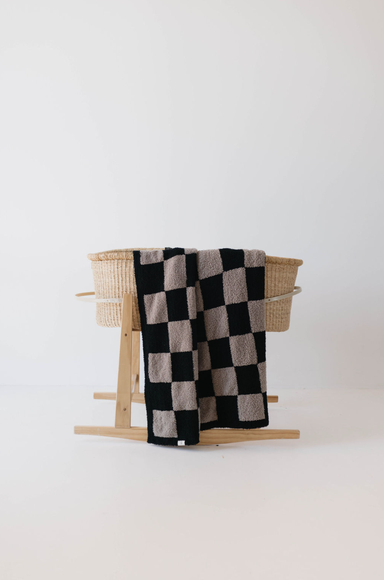 A wooden rocking stand cradles a woven bassinet. Draped over the edge is the **Plush Blanket | Checkmate Checkerboard** by **forever french baby**, featuring a soft, Oeko-tex certified fabric with a black and beige checkered pattern. The background is a plain white wall and floor, creating a minimalist and clean aesthetic.