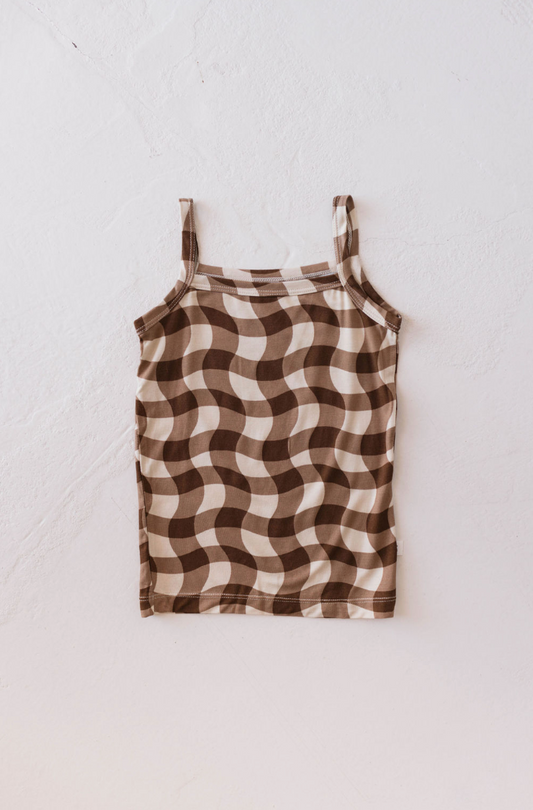 The Basic Bamboo Tank, Groovy Gingham edition by forever french baby is laid flat on a white textured surface. This sleeveless tank top, made from breathable bamboo fabric, features a beige and brown wavy checkered pattern along with thin shoulder straps and a straight neckline.