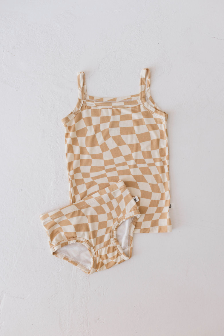 A Gold Coast Basic Bamboo Tank from forever french baby, featuring tan and white wavy, distorted checks, is paired with matching shorts and laid flat on a textured white surface. The hypo-allergenic set has a unique and playful look.