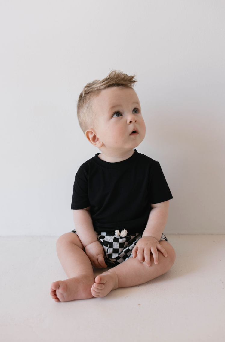 A small child with light hair sits on the floor against a plain white background, looking upwards. The child is wearing a "Basic Bamboo Tee" in Midnight Black by forever french baby and black-and-white checkered shorts. The expression on the child's face appears curious or attentive.