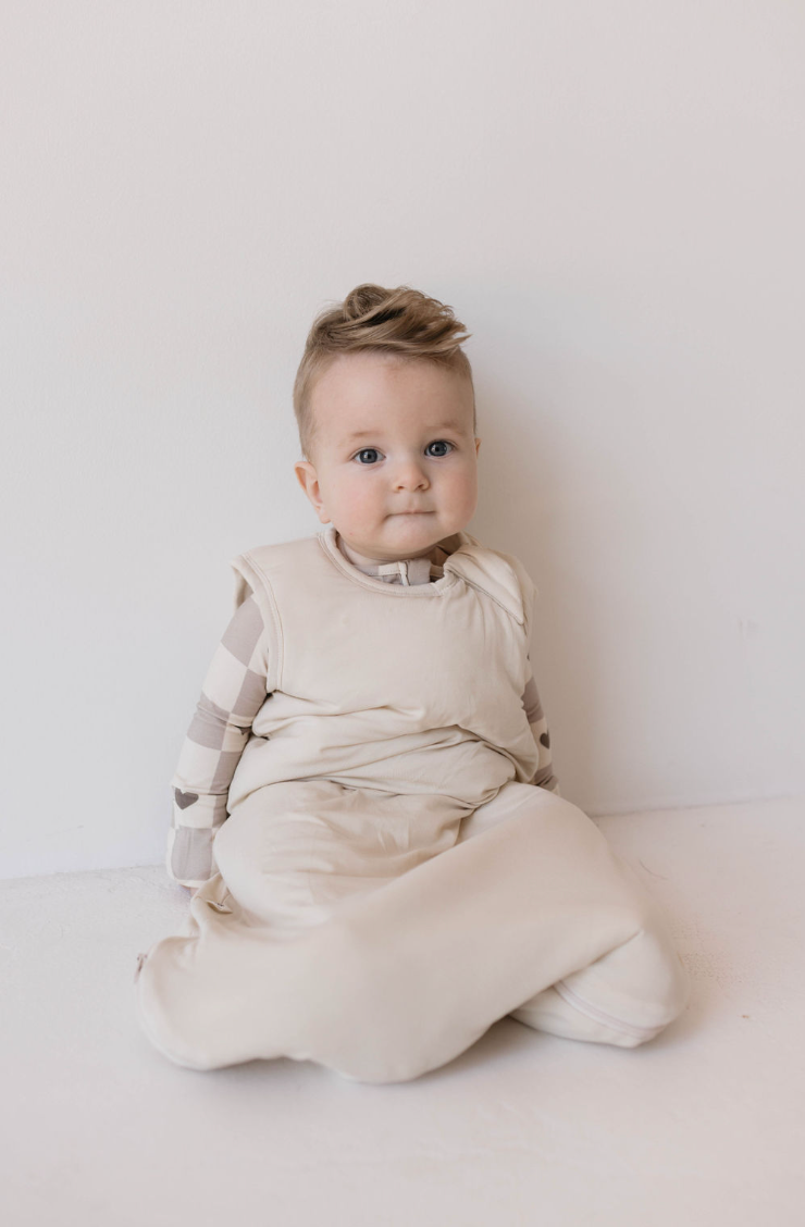 A baby wearing a "Bamboo Sleep Sack | Bone" from forever french baby with their legs tucked in and a beige and white plaid shirt sits against a plain white background, looking at the camera. The eco-friendly sleep sack complements the baby's short, light brown hair styled to the side.