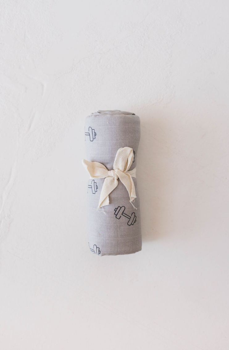 A neatly rolled Arm Day muslin swaddle in light gray, adorned with small dumbbell patterns, is secured with a cream-colored ribbon and placed on a white textured surface. Perfect for any baby's nursery, this ultra-soft blanket from forever french baby provides both comfort and style.