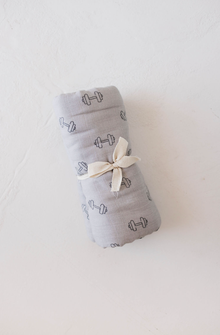 A neatly rolled Muslin Swaddle | Arm Day from forever french baby, featuring an ultra-soft fabric in light-gray adorned with a pattern of small dumbbells and tied with a cream-colored ribbon, resting on a smooth white surface.