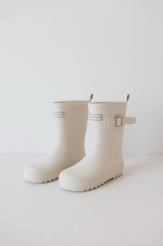 A pair of "ff Rain Boots | Cream" from the brand forever french baby are positioned side by side against a plain white background. Each boot features a small buckle detail on the side and a label on the front that reads "french." These all-weather kids' footwear boast a rugged sole for traction, showcasing their simple, minimalist design.