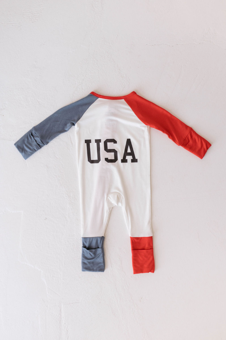 The Bamboo Zip Pajamas from Forever French Baby feature "USA" printed in large black letters on the back. These pajamas have red and blue sleeves and legs, with the left sleeve and leg in blue, and the right sleeve and leg in red. The body of the onesie is white, making it a perfect newborn outfit for celebrating the 4th of July.