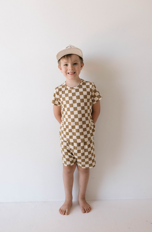 A young boy stands barefoot against a plain white background, smiling in his Bamboo Short Set | Olive Checkerboard by forever french baby. He wears a matching T-shirt and shorts in an olive checkered pattern, with a beige baseball cap on, and his hands are behind his back.