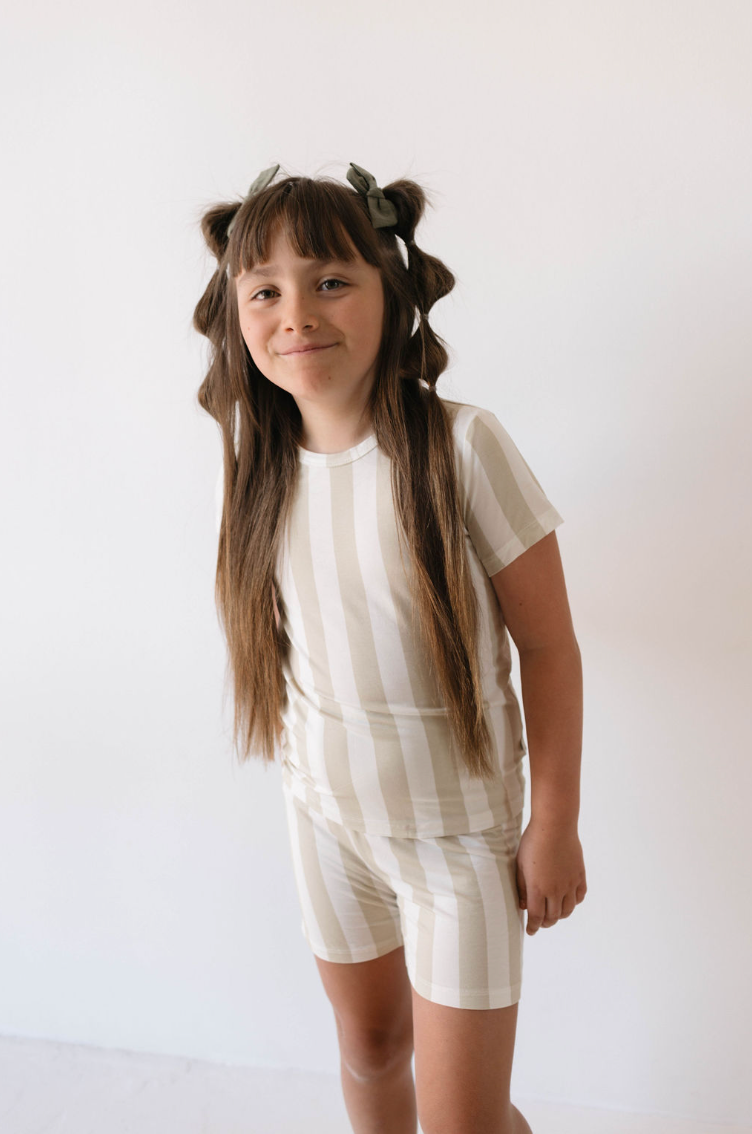 A young girl stands against a plain white background, smiling softly at the camera. She has long, straight hair styled with two green bows and wears the Bamboo Short Set in Sand Stripe by forever french baby, made from hypo-allergenic materials.