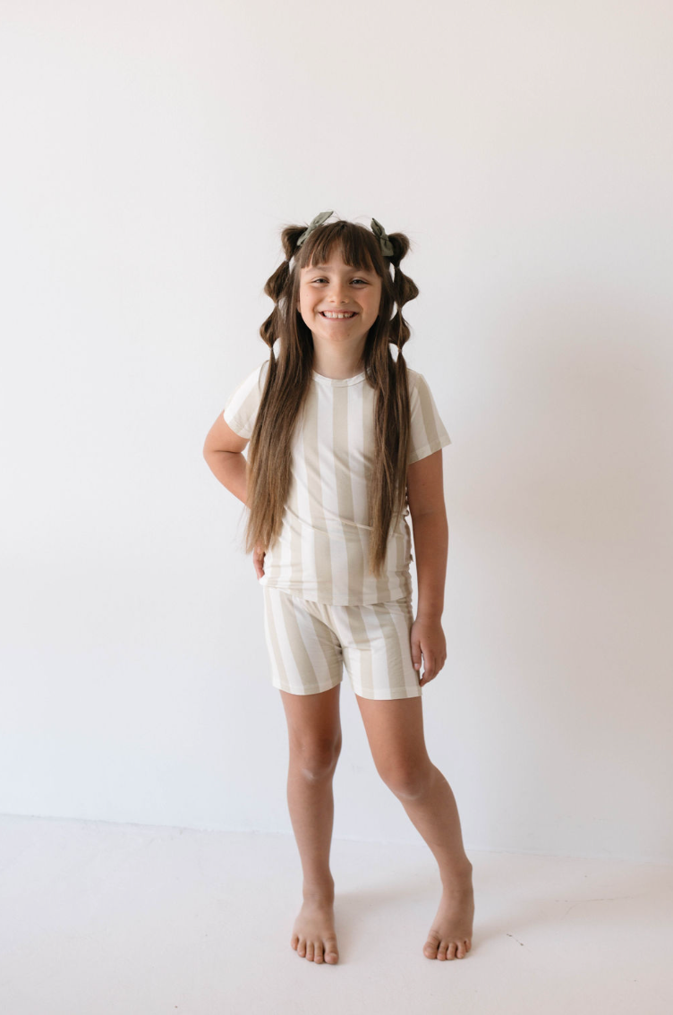 A young girl stands barefoot against a plain white background, wearing the Bamboo Short Set in Sand Stripe from forever french baby. She has long, wavy brown hair styled with bows and is smiling at the camera with her hands placed casually by her sides, showcasing hypo-allergenic clothing.