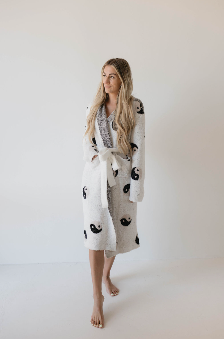 A person with long, blonde hair stands barefoot against a plain white background. They are wearing the Adult Robe | Yin Yang by Forever French, adorned with black and white yin-yang symbols and a matching white belt. The individual has a relaxed and calm expression.