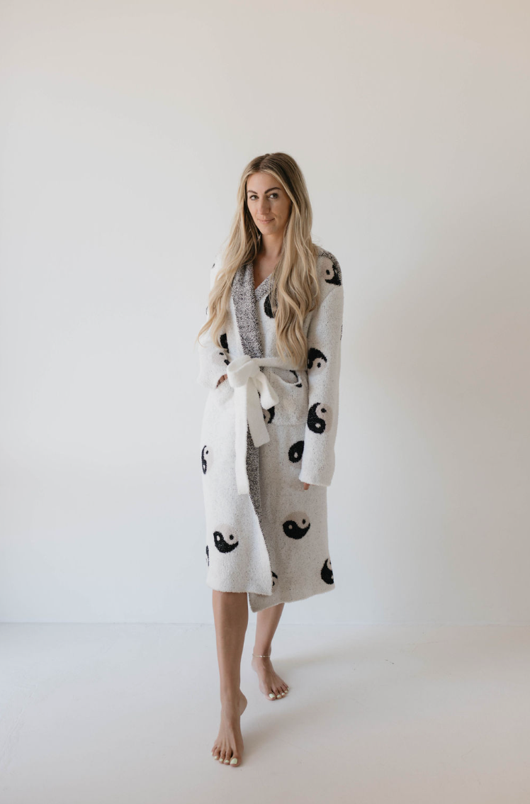 A person with long, blonde hair is standing barefoot against a plain white background. They are wearing the Adult Robe | Yin Yang by Forever French, which is OEKO-TEX certified and made of microfiber feather yarn. The robe features black yin-yang symbols and includes a tied belt at the waist. The person gazes directly at the camera with a relaxed expression.