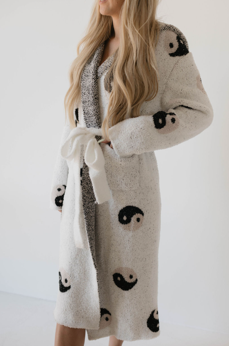 A person with long blond hair is wearing the "Adult Robe | Yin Yang" by Forever French, a white, fuzzy bathrobe made of OEKO-TEX certified microfiber feather yarn featuring black and white yin-yang symbols. The cozy robe is tied at the waist with a matching belt, and the person is standing against a plain white background facing slightly to the left.