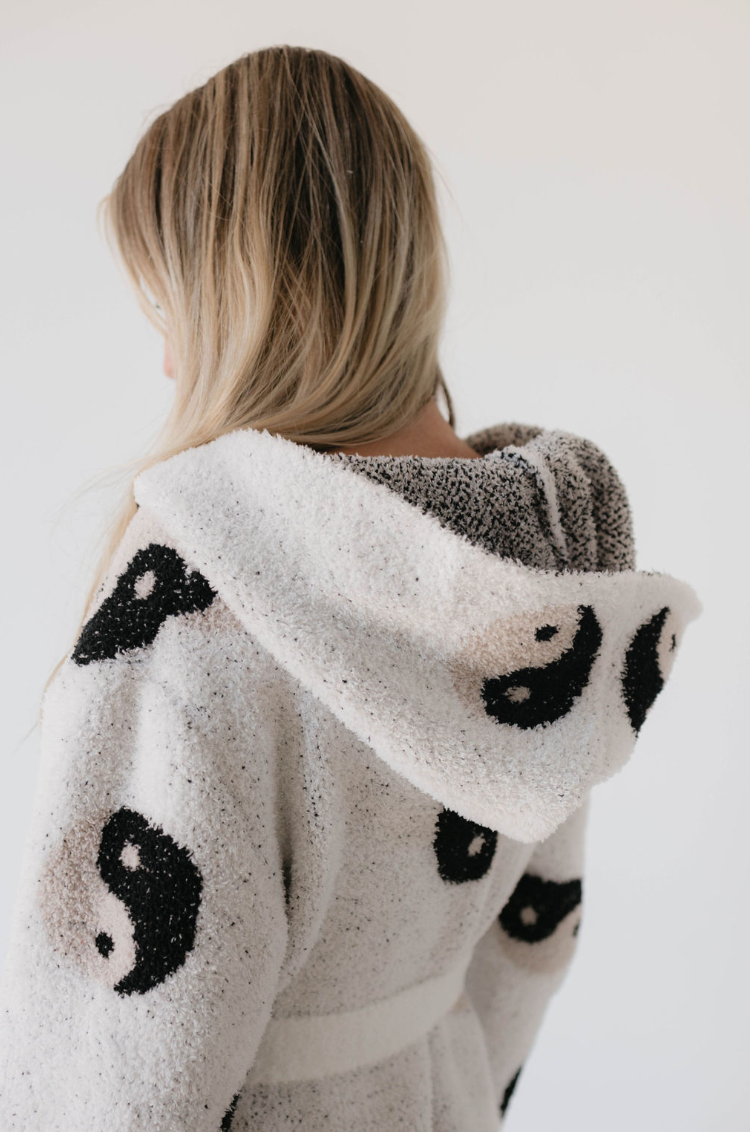 A person with long, blonde hair is seen from behind, wearing the Adult Robe | Yin Yang by Forever French. The fluffy, white robe features black yin-yang symbols scattered across it and is made from microfiber feather yarn. The OEKO-TEX certified material gives the soft robe a cozy, textured look against the plain, light-colored background.