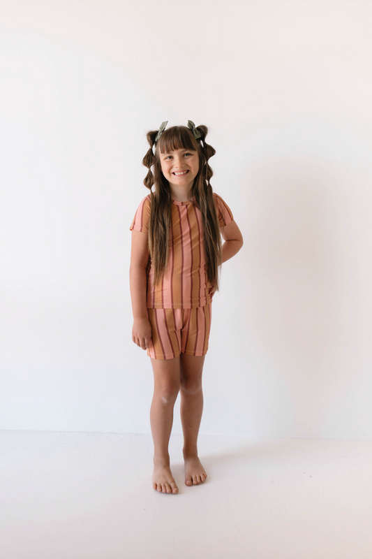 A young girl with long brown hair styled in buns and colorful ties stands barefoot smiling against a white background. She wears the Bamboo Short Sets | Boardwalk from forever french baby, featuring a pink and orange striped outfit with breathable fabric, shorts, and a short-sleeved top.