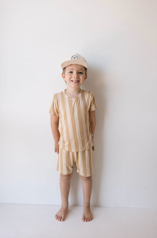 A young child stands against a white wall, smiling. They are wearing the Bamboo Short Set | Peach Stripe from forever french baby, a stylish beige and white striped outfit consisting of a short-sleeved shirt and shorts made from breathable fabric. They also have a white cap on their head and are barefoot.