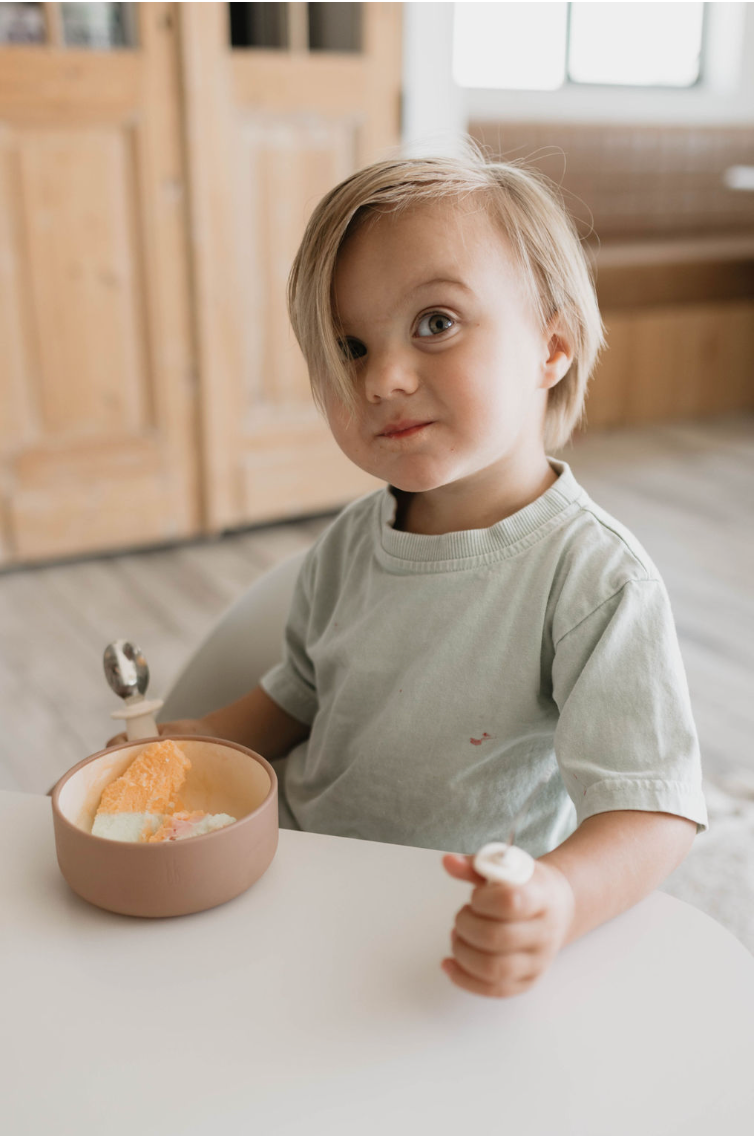 A small child with light hair sits at a table, looking at the camera with a curious expression. The child is holding a small item in one hand and has a food-safe *Silicone Bowl in Desert Brown* from *forever french baby* filled with scoops of ice cream in front of them. The background features a wooden door and a seating area.