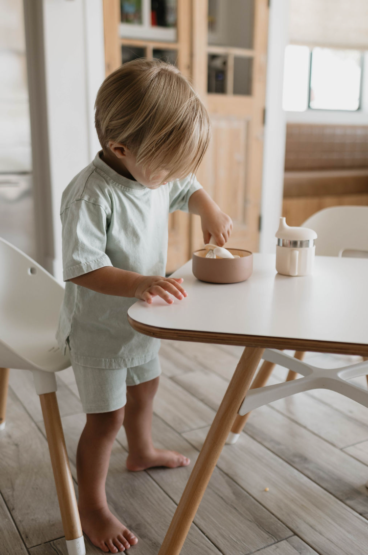 A young child with light hair stands barefoot, stirring food in a Desert Brown Silicone Bowl from forever french baby on a white table. The child is wearing a light-colored outfit consisting of a short-sleeved shirt and shorts. A cream-colored drink container sits on the table, with wooden furniture in the background.