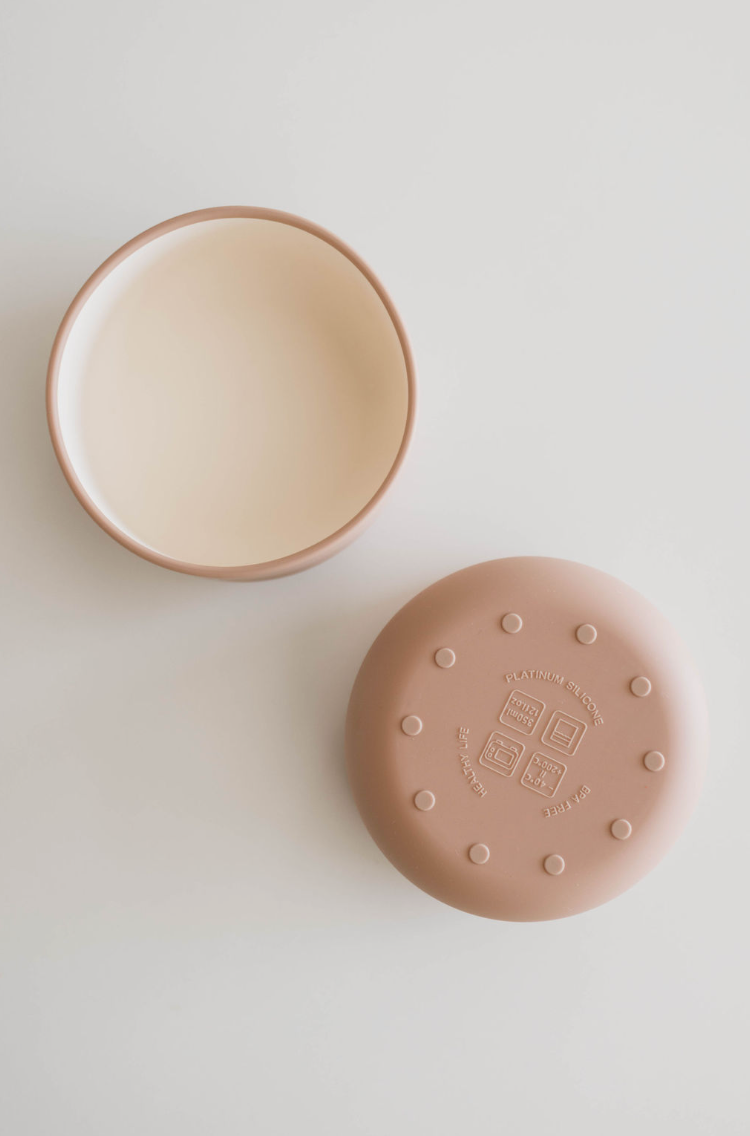 Top view of a two-piece silicone container on a white surface. The "Silicone Bowl | Desert Brown" by forever french baby features one open circular bowl with a beige interior and brown exterior, and the other is an upside-down lid, brown and round, with symbols and text embossed on it—perfect for baby and toddler meals.