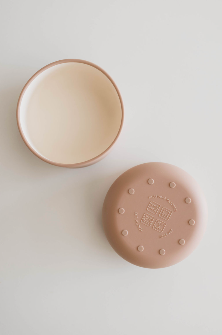 A Desert Brown, round silicone bowl by forever french baby, designed for baby and toddler meals, along with its matching lid are displayed on a white surface. The bowl is open, revealing its smooth interior, while the lid lies beside it adorned with multiple raised dots and embossed text on its food-safe silicone surface.