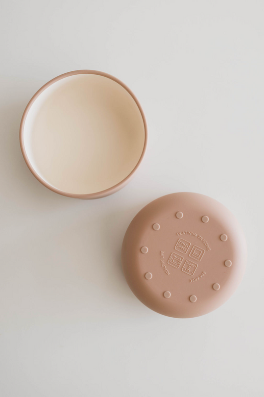 A Desert Brown, round silicone bowl by forever french baby, designed for baby and toddler meals, along with its matching lid are displayed on a white surface. The bowl is open, revealing its smooth interior, while the lid lies beside it adorned with multiple raised dots and embossed text on its food-safe silicone surface.