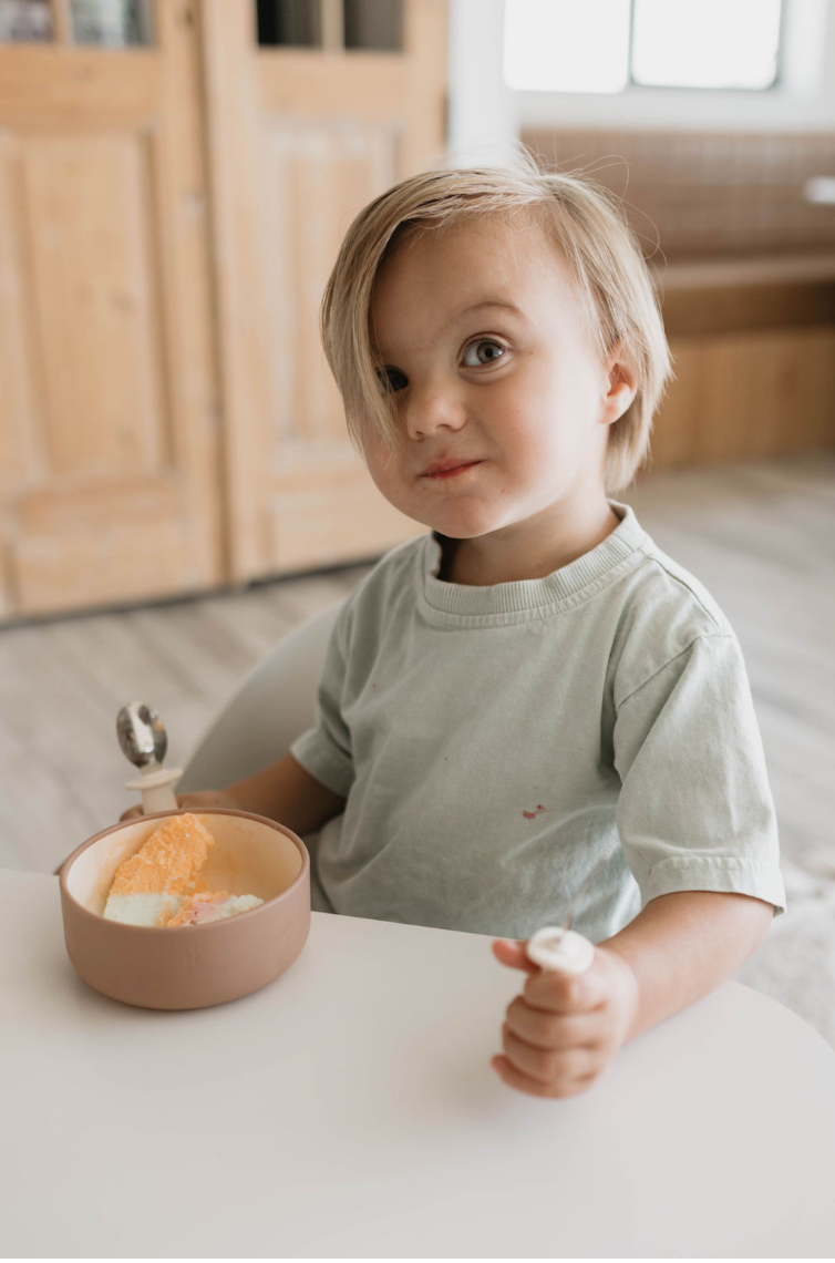 A young child with light hair sits at a table, holding a small spoon in one hand and a **forever french baby Silicone Bowl in Desert Brown** containing different colored ice creams in the other. Wearing a light green T-shirt, they look directly at the camera. Light wood furniture forms the background, highlighting this charming moment of baby and toddler meals.