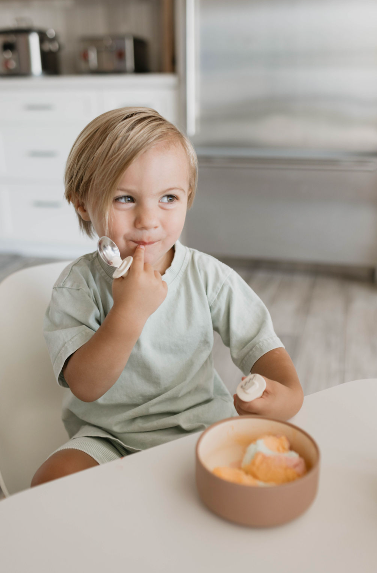 A young child with light hair, wearing a light green shirt, sits at a table in a kitchen. The child is holding a spoon and licking their finger while looking thoughtful. In front of them is a desert brown Silicone Bowl from forever french baby filled with colorful ice cream. The background features a refrigerator and cabinets.