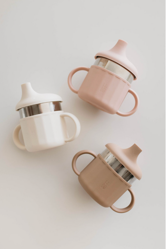 Three pastel-colored ff Sippy Cups by forever french baby are arranged on a white background. These food-safe silicone cups each feature two handles, a spout, and stainless steel interiors. The colors are soft cream, blush pink, and light brown, boasting a simple and modern design.