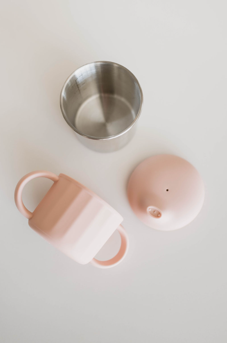 A disassembled ff Sippy Cup from forever french baby, featuring a pink exterior with two handles, an inverted dome-shaped lid, and a stainless steel inner cup, is laid out on a white surface.