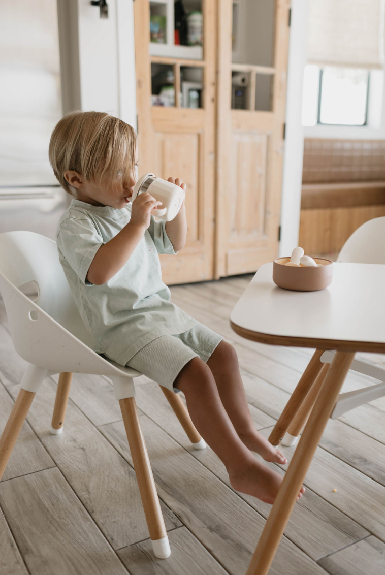 A small child with blonde hair sits on a white chair at a table in the kitchen, drinking from a forever french baby ff Sippy Cup with both hands. The child is barefoot and wearing a light green outfit. A small round bowl with eggs is on the table in front of the child.