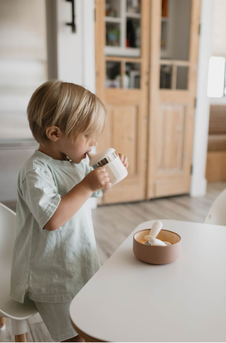 A young child with light brown hair is standing in a kitchen setting, sipping from an ff Sippy Cup by forever french baby. In front of them on the white table, there is a bowl with a spoon. The background features wooden cupboards and a stainless steel refrigerator, while the child wears a light-colored outfit.