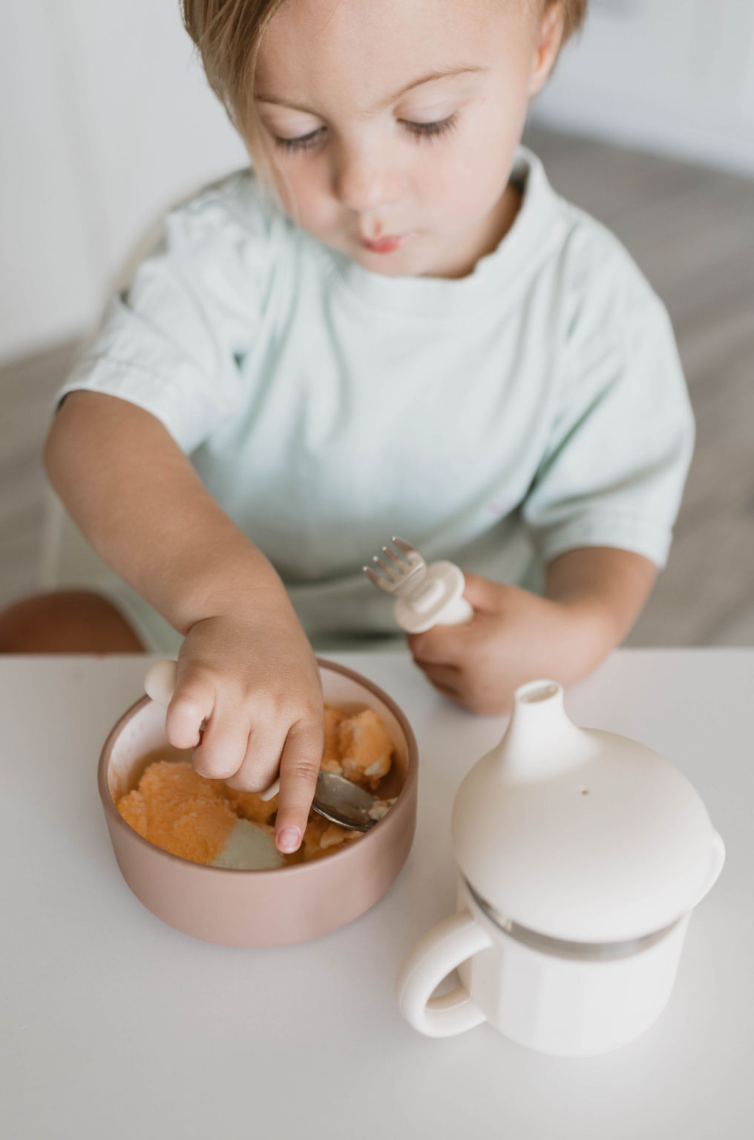 A young child with light hair sits at a table using a spoon to eat mashed food from a bowl. The child holds a stainless steel fork in the other hand, while an ff Sippy Cup from forever french baby, made of food-safe silicone and complete with a lid, is placed on the table in front of them.