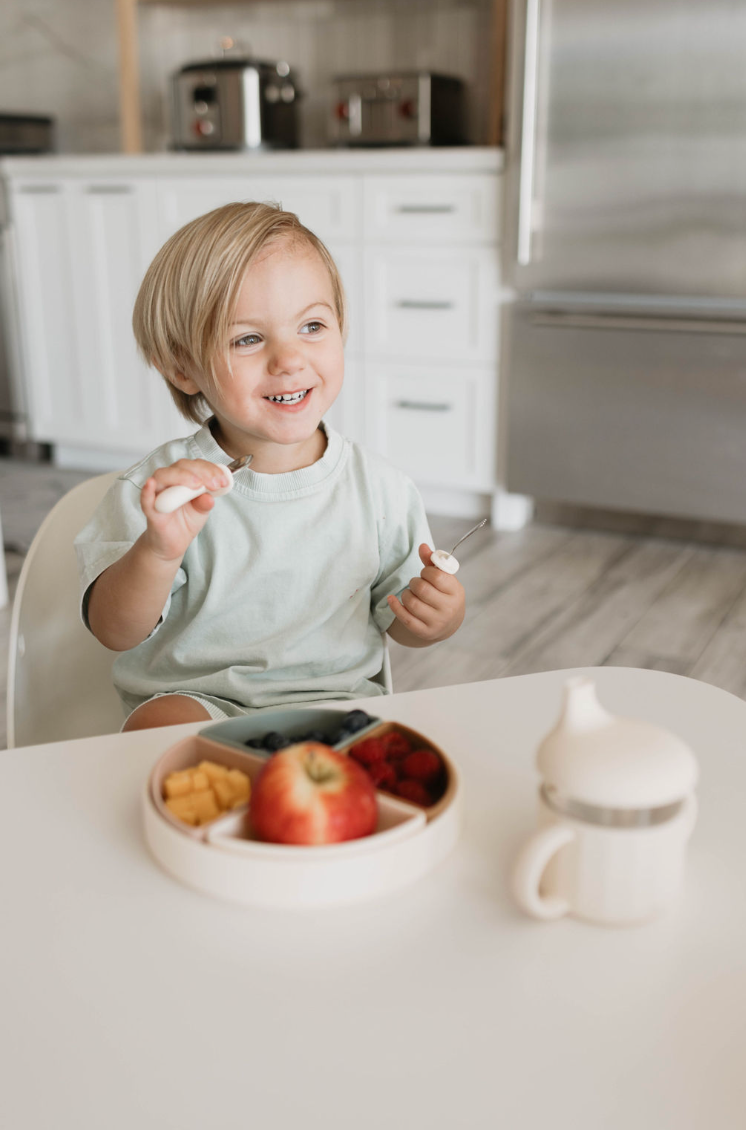 A young child with blonde hair sits at a white table in a modern kitchen during mealtime. The child is smiling and holding a fork. In front of them is the forever french baby Training Plate | Ivory, filled with various foods, including an apple and small snack pieces. A sippy cup sits nearby on the table.