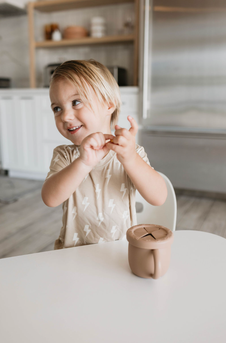 A young child with light hair, wearing a beige shirt adorned with a white lightning bolt pattern, sits at a table smiling and playing with their hands. On the table is a beige BPA-free silicone snack cup with a removable lid from forever french baby. The background features a modern kitchen with white cabinets and stainless steel appliances.