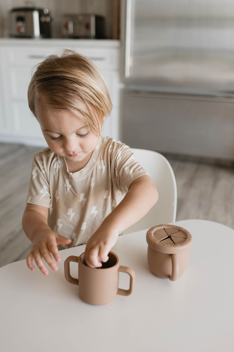 A young child with blonde hair, wearing a light-colored shirt adorned with a lightning bolt pattern, is sitting at a white table. The child is playing with two beige-colored "Snack Cup with Removable Lid" products by forever french baby in a modern kitchen featuring stainless steel appliances in the background.