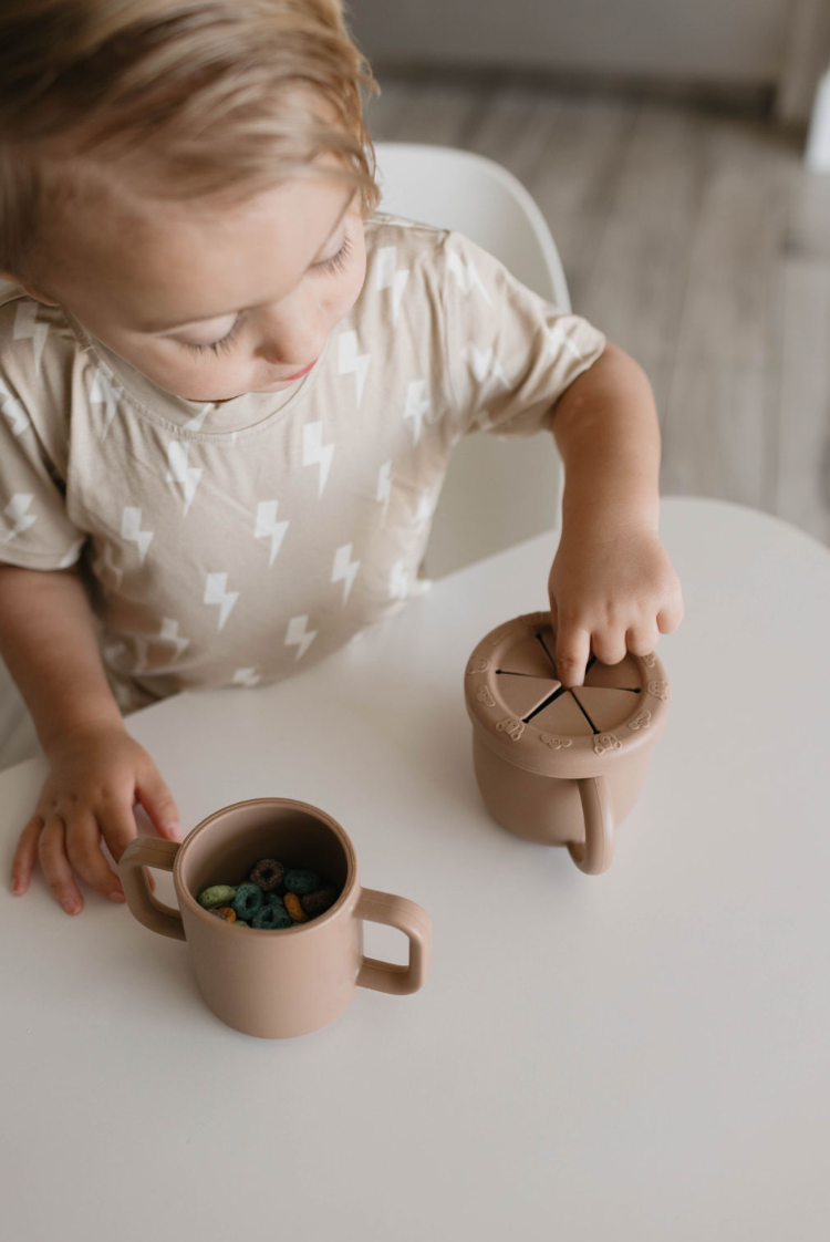 A small child with light hair sits at a white table, wearing a beige shirt with a lightning bolt pattern. The child is placing their fingers into the easy snacking BPA-free silicone Snack Cup with Removable Lid from forever french baby, which is next to a two-handled cup full of colorful cereal pieces.