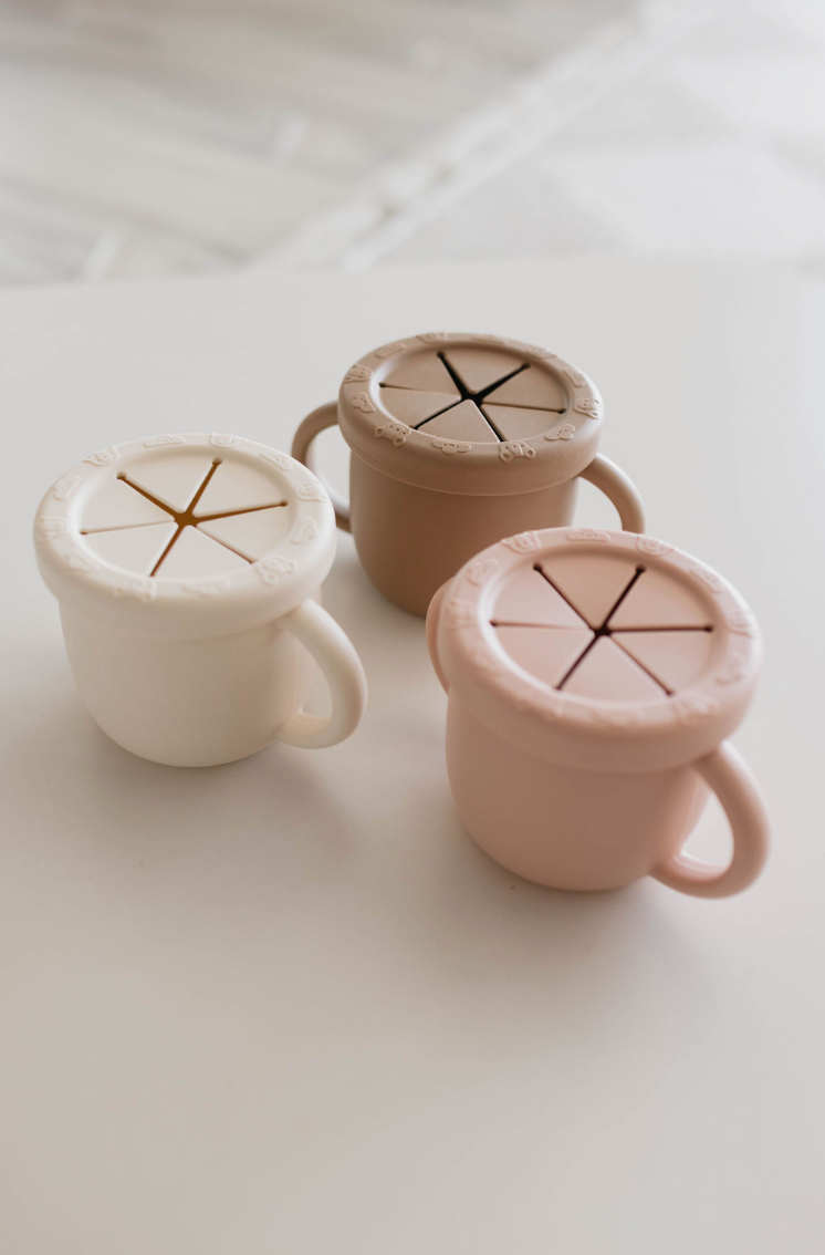 Three forever french baby Snack Cups with Removable Lids, made from BPA-free silicone and available in pastel colors (white, beige, and pink), are arranged on a white surface. Each cup features a handle and a slit lid designed for easy snacking.